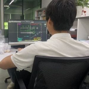 A person with short dark hair, wearing a white polo shirt, sits at a desk meticulously crafting a pogo pin 2D model on CAD software displayed on a large monitor. The office space boasts shelves and potted plants, framing the screens intricate technical drawings.