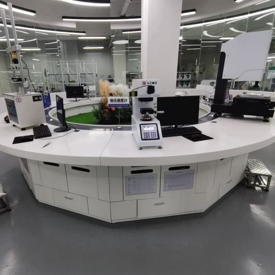 A modern laboratory serves as a quality control room with a circular white workstation equipped with computers and scientific instruments. Glass windows reveal other lab areas. Lush plants add greenery to the background. The grey floor complements the sleek setting, ensuring precision like a pogo pin connection.
